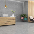 Das Bild zeigt einen modernen Empfangsbereich mit eleganten grauen Fliesen, die sowohl auf dem Boden als auch an der Wand verlegt sind. Die Fliesen haben eine dezente Marmorstruktur, die dem Raum eine stilvolle und ruhige Atmosphäre verleiht. Der Empfangstresen aus Holz mit dem "RECEPTION"-Schild ist schlicht und funktional, während die goldenen Pendelleuchten und die grünen Pflanzen einen schönen Farbakzent setzen. Die Wand im Hintergrund aus Holzpaneelen fügt Wärme hinzu und kontrastiert angenehm mit den kühlen grauen Fliesen und Möbeln. Diese Gestaltung sorgt für eine einladende, moderne und professionelle Ausstrahlung im Raum. Wenn du mehr Informationen zu den Fliesen oder zur Raumgestaltung benötigst, stehe ich dir gerne zur Verfügung!